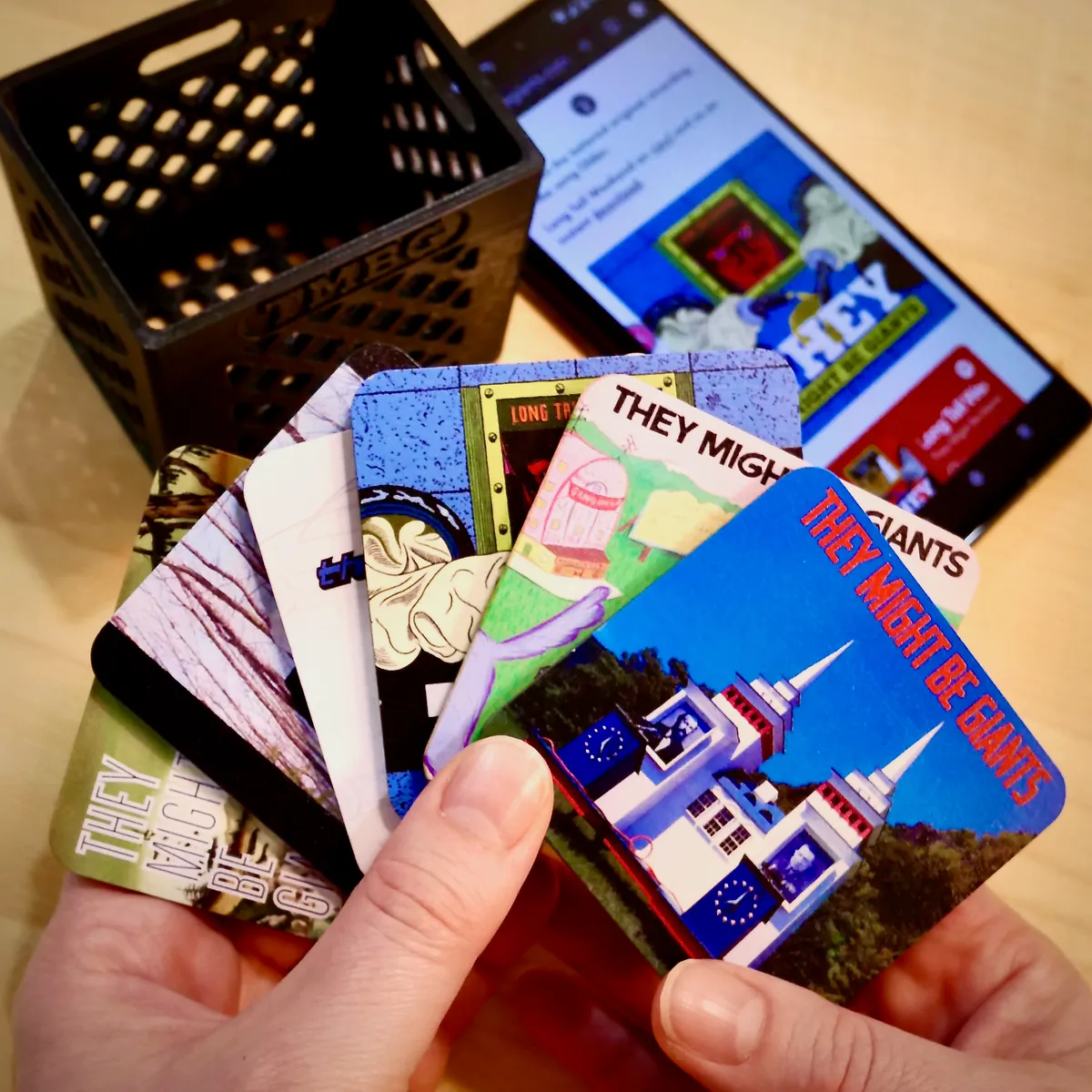A fan of Goodz cards held in two hands, with a wooden crate of Goodz and a phone showing a Bandcamp page in the background.